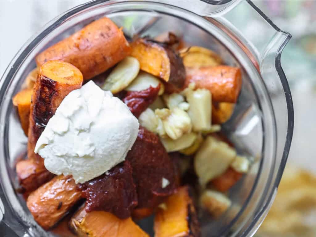 Close-up of a blender filled with roasted sweet potatoes, gochujang, garlic cloves, and a dollop of silken tofu, ready to be blended.